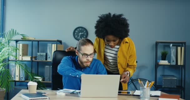 Ocupado hombre y mujer de raza mixta en la oficina. Caucásico hombre escribiendo en el ordenador portátil mientras está sentado en el escritorio y mostrando algo a su jefe femenino. chica afroamericana mirando a la pantalla de la computadora mientras está de pie — Vídeo de stock