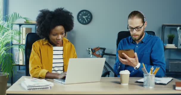 Hombre y mujer de raza mixta trabajando en la oficina. Hombre caucásico en gafas golpeando en la tableta y mostrando a colega mujer. Chica afroamericana sentada en el escritorio y mensajes de texto en el portátil. Concepto de trabajo — Vídeo de stock