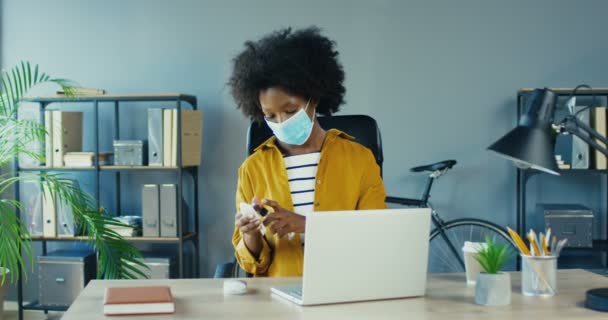 Γυναίκα εργαζόμενος απολύμανση ποντίκι υπολογιστή και σημειωματάριο στο γραφείο σε καραντίνα. Όμορφη Αφροαμερικανή γυναίκα σε μάσκα καθαρισμού χώρο εργασίας με αντισηπτικό, ενώ κάθεται. Έννοια προστατευτικών μέτρων — Αρχείο Βίντεο