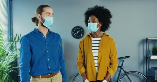 Fröhliche multiethnische Mitarbeiter in medizinischen Masken stehen im Büro und schauen einander an. Afroamerikanerin schöne Frau in guter Laune. Schöner Kaukasier, der in die Kamera blickt. Quarantäne-Konzept — Stockvideo