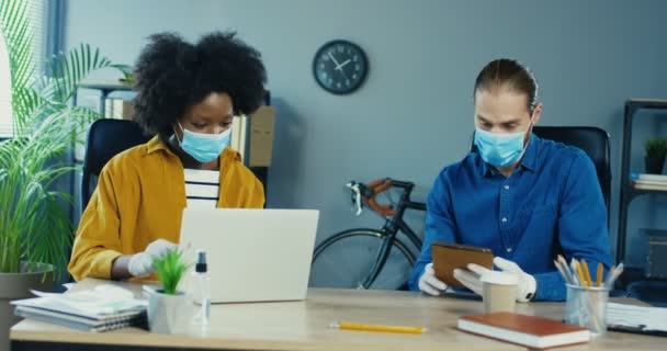 Wieloetniczni pracownicy w maskach i rękawiczkach pracujący w gabinecie. Dziewczyna pisząca na laptopie. biały przystojny mężczyzna stukanie na tablet i pokazując dla piękny Afroamerykanka podczas siedzi w biuro — Wideo stockowe