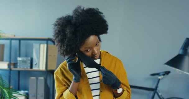 Портрет красивої афро-американської дівчини в рукавичках, одягненої захисної маски на обличчі в офісі в приміщенні. Красуня-працівниця в кабінеті позує перед камерою. Працюю в карантині. Завершальна концепція — стокове відео