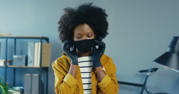 Primer plano retrato de una mujer afroamericana bastante alegre posponiendo la máscara protectora en el interior del gabinete. Hermosa empleada en el lugar de trabajo sonriendo a la cámara. Trabaja en cuarentena. Concepto de trabajador — Vídeos de Stock