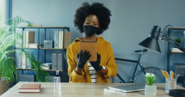 Koruyucu maskeli ve eldivenli güzel bayan ofis çalışanı karantinadaki tabletlere bakıyor ve onları inceliyor. Güzel Afro-Amerikalı kadın iş yerinde bir cihaz kullanıyor. Görev konsepti