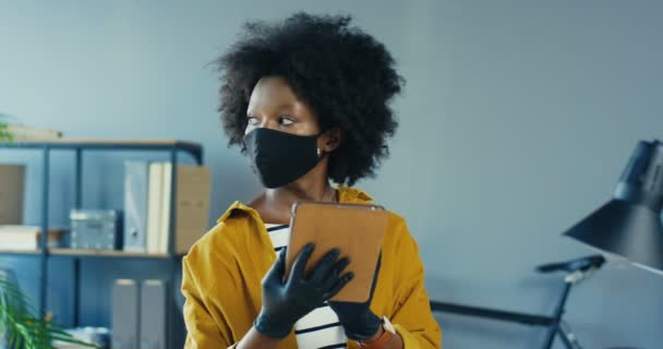Güzel bayan ofis çalışanı gözlerini kaçırıyor ve kapalı dolaptaki tablete bakıyor. İş yerindeki karantina konseptinde koruyucu maske takan güzel Afrikalı Amerikalı kızın portresini kapat. — Stok video