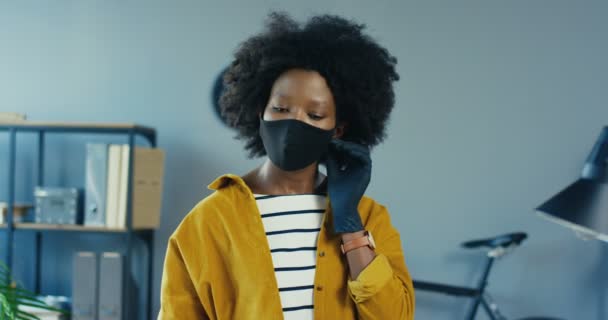 Primer plano retrato de hermosa niña afroamericana alegre posponer la máscara protectora en la oficina en el interior. Bastante empleada en el gabinete sonriendo a la cámara. Trabaja en cuarentena. Concepto de trabajo — Vídeos de Stock