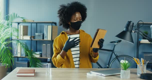 Jolie femme d'affaires afro-américaine en masque de protection et gants vidéochat sur tablette au bureau. Belle travailleuse ayant un appel vidéo alors qu'elle était assise dans le placard. Concept de travail — Video