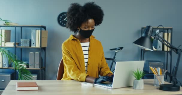 Mujer de negocios bastante afroamericana con máscara protectora y guantes trabajando en la computadora mientras se sienta en el escritorio en la oficina y sonríe a la cámara. Hermosa trabajadora escribiendo en el portátil. Concepto de gabinete — Vídeos de Stock