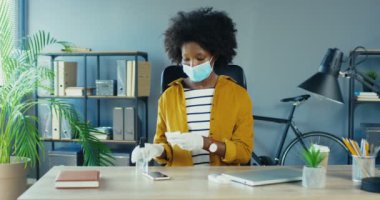 Tıbbi maskeli ve eldivenli Afro-Amerikan kadın karantinada ofisinde oturuyor ve akıllı telefonu dezenfektanla dezenfekte ediyor. Dolabın masasında güzel bir bayan işçi var. Güvenlik önlemleri kavramı