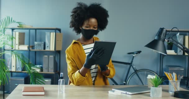 Koruyucu maskeli ve eldivenli güzel, ciddi bir bayan çalışan karantina ofisindeki tablette geziniyor. Güzel Afrikalı Amerikalı kadın iş yerindeki dolapta cihaza dokunmakla meşgul. Görev konsepti — Stok video