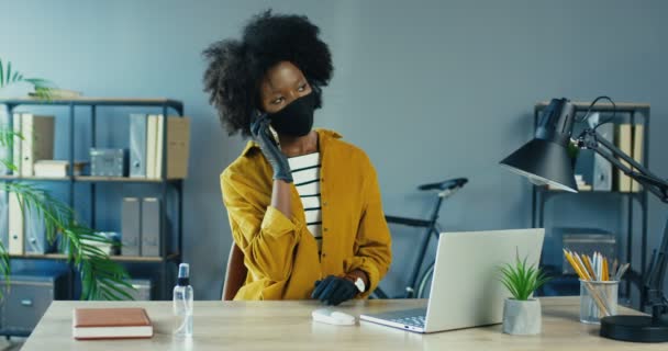 Yüz maskeli ve eldivenli güzel Afro-Amerikan iş kadını iş yerinde otururken akıllı telefondan arıyor. Ofiste cep telefonuyla konuşan oldukça meşgul bir bayan çalışan. Karantina konsepti — Stok video