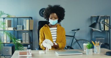 Tıbbi maskeli güzel Afro-Amerikan kadın eldiven takıyor ve ofis masasında otururken dizüstü bilgisayarını açıyor. Karantina istihdam kavramında bilgisayar üzerinde çalışırken daktilo yazan güzel bir bayan işçi.