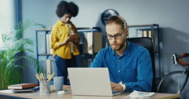 Ofis masasında otururken laptopta yazı yazan yakışıklı, beyaz, ciddi bir adam. Güzel Afrikalı Amerikalı meşgul kadın arka planda duruyor ve tableti tıkırdatıyor. Çok ırklı çalışanlar. Çalışma konsepti