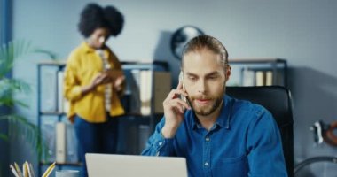 Ofis masasında otururken akıllı telefondan konuşan yakışıklı beyaz adamı yakından çek. Afro-Amerikalı kadın arka planda duruyor ve tablete vuruyor. Çok ırklı çalışanlar iş konsepti