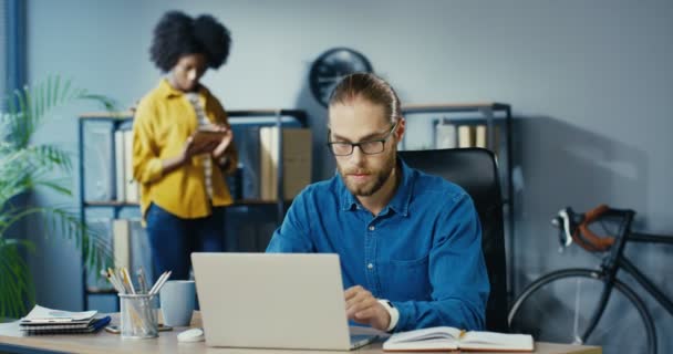 Bellissimo lavoratore caucasico di sesso maschile che digita sul computer portatile e scrive nel quaderno mentre è seduto in ufficio. Ragazza afro-americana che cammina sullo sfondo e picchietta sul tablet nel gabinetto. Concetto di razza mista — Video Stock