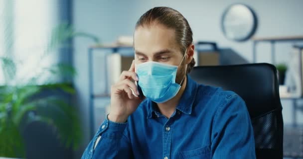 Bello giovane impiegato maschile in maschera medica che parla sullo smartphone mentre è seduto nell'armadietto in isolamento. dipendente caucasico avendo conversazione sul cellulare sul posto di lavoro Close up concetto ritratto — Video Stock