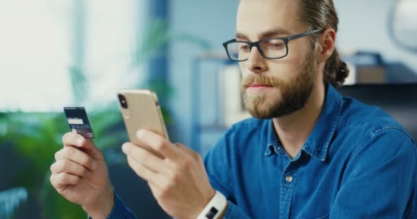 Nahaufnahme Porträt eines gut aussehenden kaukasischen Typen, der online auf dem Smartphone kauft, während er im Kabinett sitzt. Fröhlicher Mann mit Brille bezahlt mit Kreditkarte beim Internet-Einkauf per Handy — Stockvideo