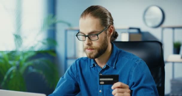 Primer plano retrato de hombre caucásico guapo feliz de compras en línea en el ordenador portátil mientras está sentado en el gabinete. Hombre alegre en gafas que pagan con tarjeta de crédito mientras compra en Internet en el lugar de trabajo Concepto de comprador — Vídeos de Stock