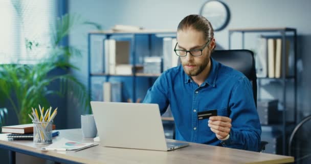 Vrolijke knappe blanke man met bril online winkelen op laptop terwijl hij in de kast zit. Vreugdevolle tevreden man betalen met credit card tijdens het kopen op internet op de werkplek. Shopaholic-concept — Stockvideo