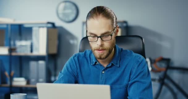 Close up retrato de homem barbudo bonito em óculos trabalhando no laptop e olhando para longe no armário. Caucasiano homem de negócios sério sentado no local de trabalho dentro de casa e usando computador. Conceito de emprego — Vídeo de Stock