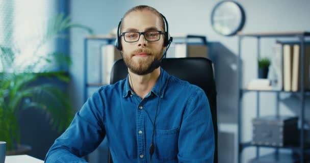 Portrét pohledného pracovníka kavkazské kanceláře ve sluchátkách, který se usmívá do kamery, zatímco sedí za stolem ve skříni. Veselý muž v brýlích v dobré náladě na pracovišti. Koncept zaměstnanosti — Stock video