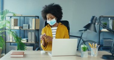 Tıbbi maskeli güzel bir bayan çalışan ellerini dezenfekte ediyor ve karantinada bilgisayar üzerinde çalışıyor. Ofis masasında otururken dizüstü bilgisayarda yazan güzel Afro-Amerikan kadın. Karantina konsepti