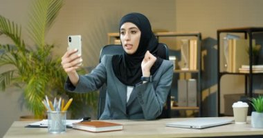 Başörtülü güzel Müslüman kadın ofiste masada oturuyor ve akıllı telefondan webcam ile konuşuyor. Güzel Arap iş kadını tesettürlü cep telefonunda video kaset taşıyor. Video sohbeti.