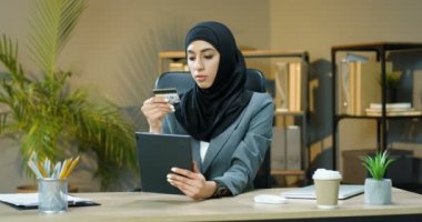 Geleneksel tesettürlü genç Müslüman kadın tablet cihazıyla online alışveriş yapıyor ve ofiste otururken kredi kartıyla ödüyor. İnternetten internet alan güzel bir Arap kadın..