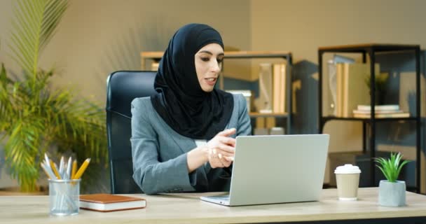 Piękna muzułmańska młoda kobieta w tradycyjnej chustce siedzi przy stole w biurze i rozmawia przez kamerę internetową na laptopie. Piękna arabska bizneswoman w hidżabie ma video na komputerze. Wideokatowanie. — Wideo stockowe