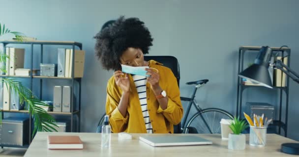 Krásná Afroameričanka si nasazuje lékařskou masku a dezinfikuje si ruce, zatímco sedí u psacího stolu. Pěkná zaměstnankyně otevírá notebook a pracuje na počítači v karanténě. Koncept úlohy — Stock video