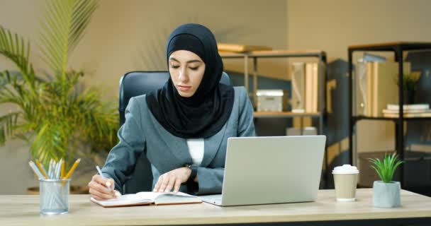 Joven musulmana hermosa mujer en hijab tradicional sentado en el escritorio en la oficina frente a la computadora portátil y la escritura en el cuaderno. Mujer de negocios en pañuelo para la cabeza notando en planificador. Concepto de planificación empresarial — Vídeos de Stock