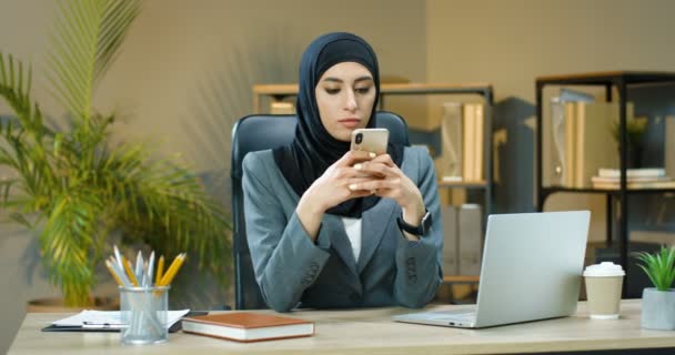 Arabian όμορφη νεαρή γυναίκα σε hijab κάθεται στο τραπέζι στο άνετο γραφείο και χτυπώντας ή κύλιση σε smartphone μπροστά από φορητό υπολογιστή. Μουσουλμάνα επιχειρηματίας γραπτό μήνυμα στο κινητό τηλέφωνο. — Αρχείο Βίντεο