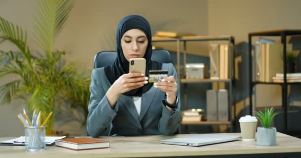 Mujer musulmana joven bastante feliz en las compras tradicionales de pañuelos para la cabeza en línea en el teléfono inteligente y pagar con tarjeta de crédito mientras está sentado en el escritorio en la oficina. Hermosa compra femenina árabe en internet por teléfono — Vídeos de Stock