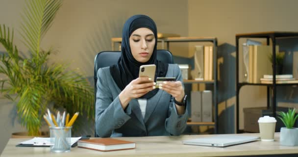 Симпатичная довольная молодая мусульманка в головном уборе покупает онлайн-платки на смартфоне и платит кредитной картой, сидя за столом в офисе. Арабская женщина покупает в интернете по мобильному телефону. — стоковое видео