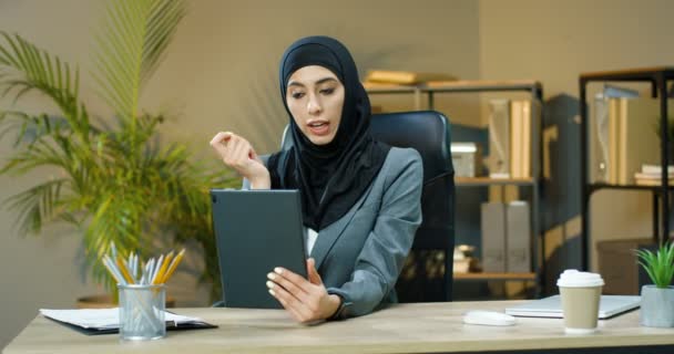 Schöne muslimische junge Frau mit Kopftuch sitzt am Tisch im Büro und spricht via Webcam auf Tablet-Gerät. Hübsche arabische Geschäftsfrau im Hidschab mit Videochat über Gadget. Videochat-Computer. — Stockvideo