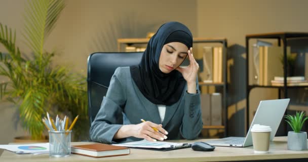 Mujer árabe cansada en hijab sentado en el escritorio en la oficina, pensando y trabajando en el ordenador portátil. Administradora musulmana escribiendo en la computadora y anotando en documentos con gráficos y gráficos. Mujer de negocios árabe. — Vídeos de Stock