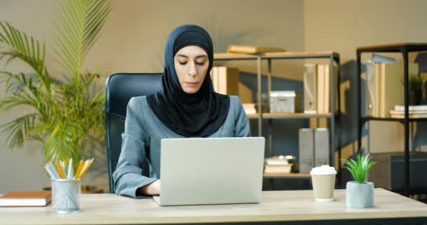 Przystojna, młoda Arabka w hidżabskim szaliku, siedząca przy biurku i pracująca na laptopie. Menedżerka muzułmańska wpisująca na klawiaturze komputera przy stole w szafce. Arabska bizneswoman. — Wideo stockowe