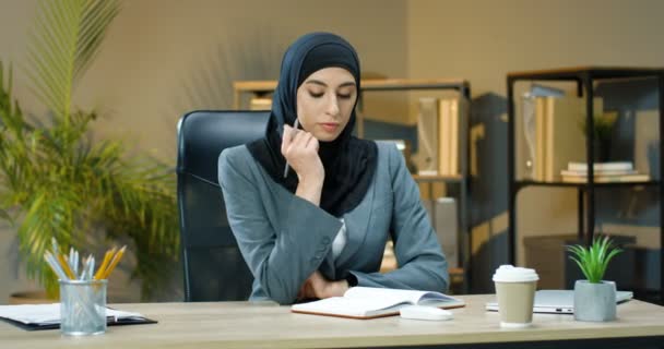 Porträt einer schönen muslimischen Frau mit traditionellem schwarzem Kopftuch, die mit Notizbuch am Tisch im Büro sitzt und in die Kamera blickt. Arabische Geschäftsfrau mit Lächeln am Schreibtisch mit Planer — Stockvideo