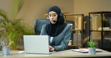 Siyah tesettürlü ve kulaklıklı genç müslüman kadın ofiste müşterisiyle bilgisayar başında web kamerasıyla konuşuyor. Arabistanlı çağrı sansür memuru bayan yönetici video şapkalı müşteriyle konuşuyor. Video sohbeti.