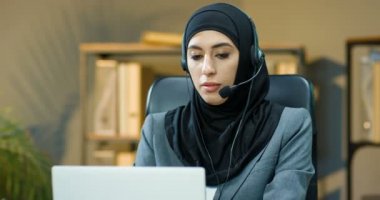 Siyah başörtüsü ve kulaklık takmış genç Müslüman kadın ofiste müşteriyle birlikte laptop bilgisayarında web kamerasıyla konuşuyor. Arap telefon sansürcüsü bayan yönetici video kasette müşteriyle konuşuyor. Video sohbeti.