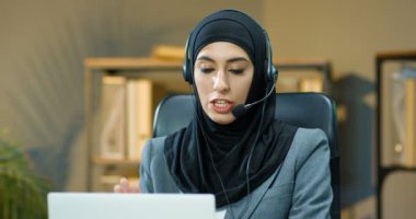 Başörtüsü ve kulaklık takmış genç mutlu müslüman kadın portresi ofiste müşteriyle birlikte laptop bilgisayarında webcam ile konuşuyor. Arapça çağrı sansür memuru bayan yönetici video kaset ve gülümseme müşteri ile konuşuyor.