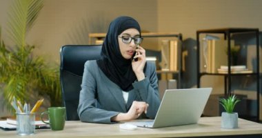 Tesettürlü ve gözlüklü genç bir Arap kadın ofisinde oturuyor, cep telefonuyla konuşuyor ve dizüstü bilgisayarla çalışıyor. Bilgisayarın klavyesinde yazan Müslüman kadın, cep telefonuyla konuşan Arap iş kadını.
