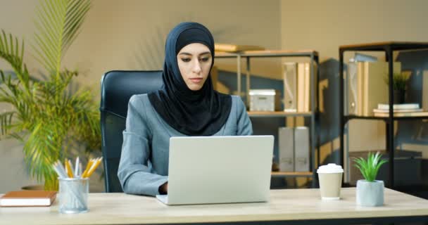 Beautiful young Arabik woman in traditional headscarf sitting at table in cozy office and working on laptop. Female muslim worker typing on keyboard of computer at desk. Arab businesswoman. — Stock Video