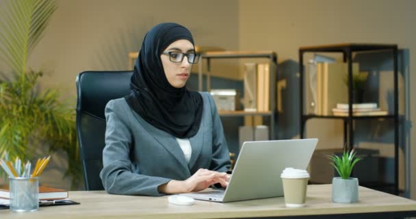 Portret młodej Arabki w hidżabie i okulary siedzącej przy stole w biurze, pracującej na laptopie i uśmiechniętej do kamery. Muzułmanka wpisująca na klawiaturze komputera przy biurku i uśmiechająca się. — Wideo stockowe