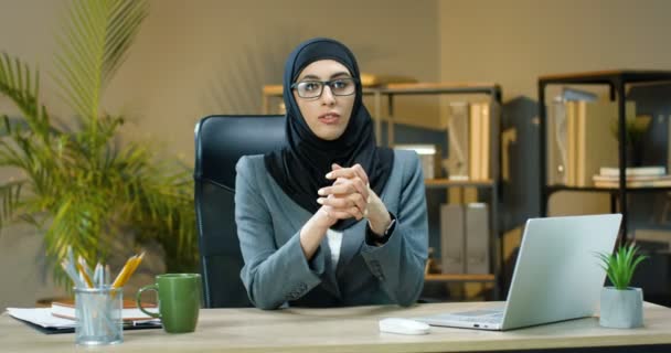 Joven mujer musulmana hermosa en hijab y gafas sentadas en la mesa en la oficina y hablando con la cámara como diciendo algo. Vlogger mujer árabe de negocios. Seminario en línea. Habla la empresaria. Vlog — Vídeo de stock