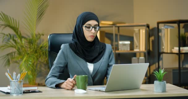 Mujer árabe joven en hijab y gafas sentadas en el escritorio en la oficina y trabajando en el ordenador portátil. Muslim hembra escribiendo en el teclado de la computadora y sorbiendo bebida caliente en la mesa en el gabinete. Beber café o té. — Vídeos de Stock