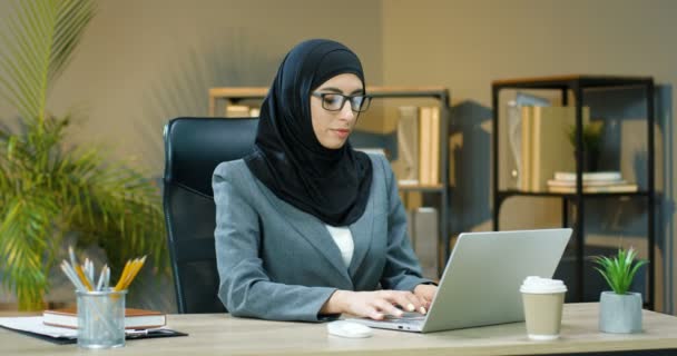 Hermosa mujer árabe joven en hijab negro y gafas sentadas en el escritorio en la oficina y trabajando en el ordenador portátil. Administradora musulmana escribiendo en el teclado de la computadora en la mesa. Café para llevar. Mujer de negocios árabe — Vídeo de stock