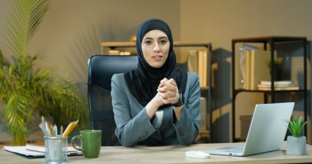 Hermosa mujer musulmana en hijab sentada en el escritorio en la oficina y hablando con la cámara como diciendo y enseñando. Blogger mujer árabe de negocios. Seminario en línea. Mujer de negocios hablando y grabando vlog — Vídeos de Stock