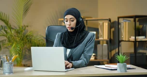 Junge muslimische Frau in schwarzem Hijab und Kopfhörer, die sich per Webcam am Laptop mit dem Kunden im Büro unterhält. Arabische Callcenter-Managerin spricht im Videochat mit Kunden. Videochat. — Stockvideo