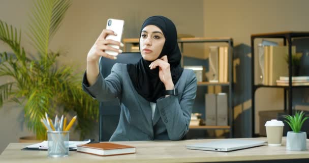 Hermosa joven musulmana en hijab tradicional sentado en el escritorio en la oficina y posando a la cámara del teléfono inteligente. Mujer árabe de negocios tomando foto selfie en el teléfono móvil. En el interior. Trabajadora empresarial. — Vídeos de Stock